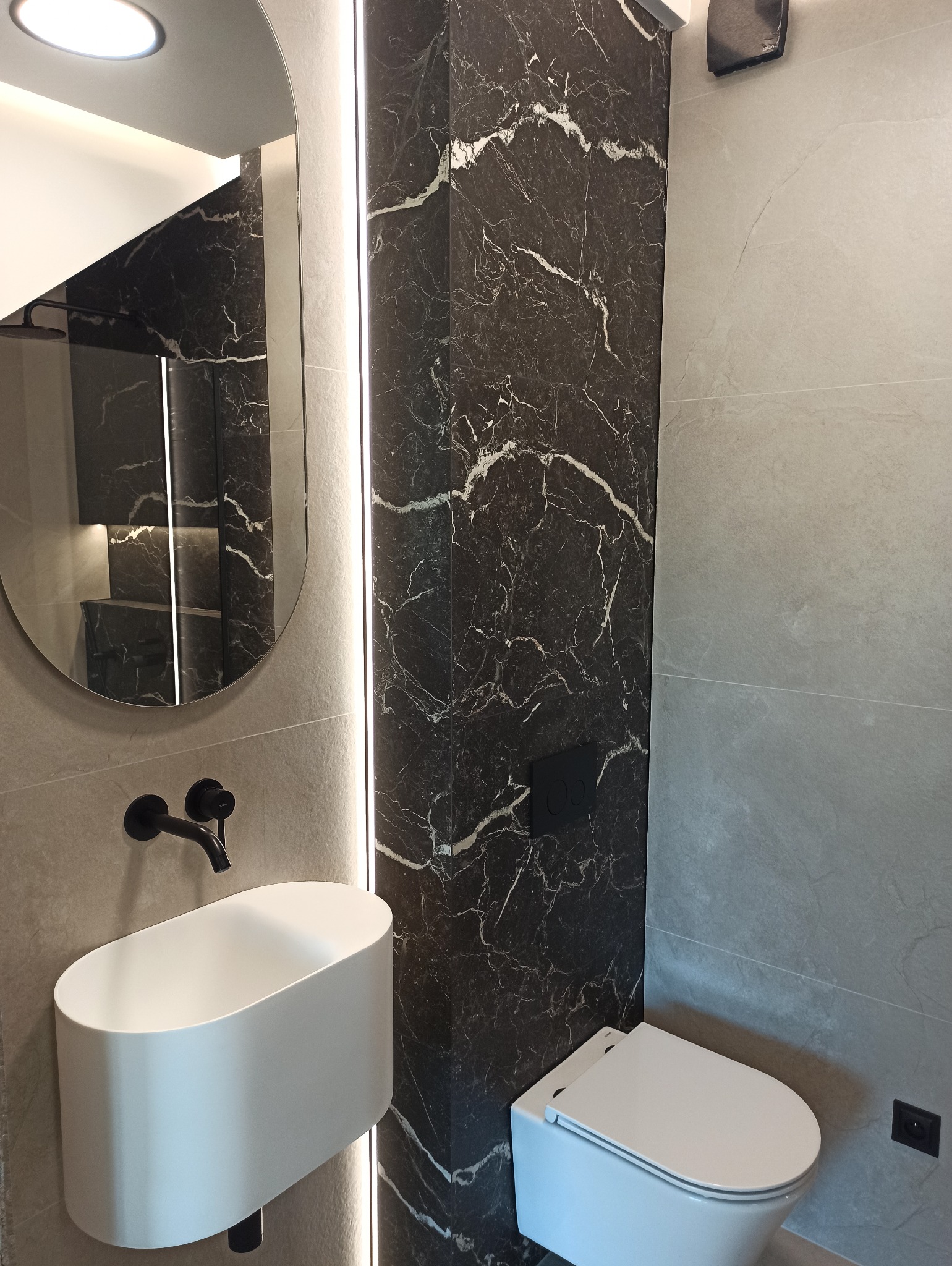 Nowoczesna łazienka z owalnym lustrem z podświetleniem LED, umywalką nablatową i czarną płytką imitującą marmur. Minimalistyczny design.