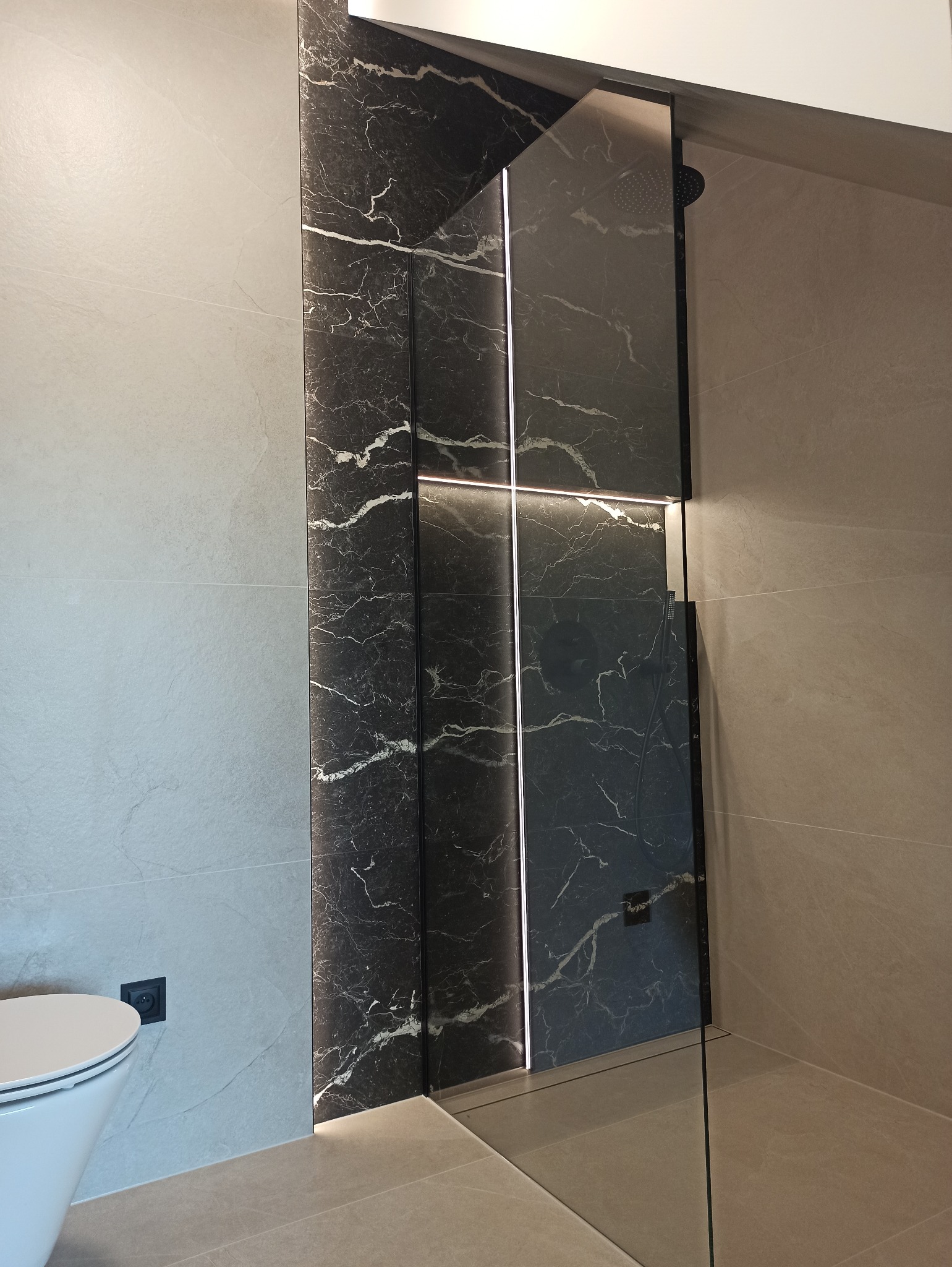 Nowoczesna kabina prysznicowa z czarnymi płytkami imitującymi marmur, podświetlona listwą LED, na tle beżowych płytek. Widoczny fragment toalety.