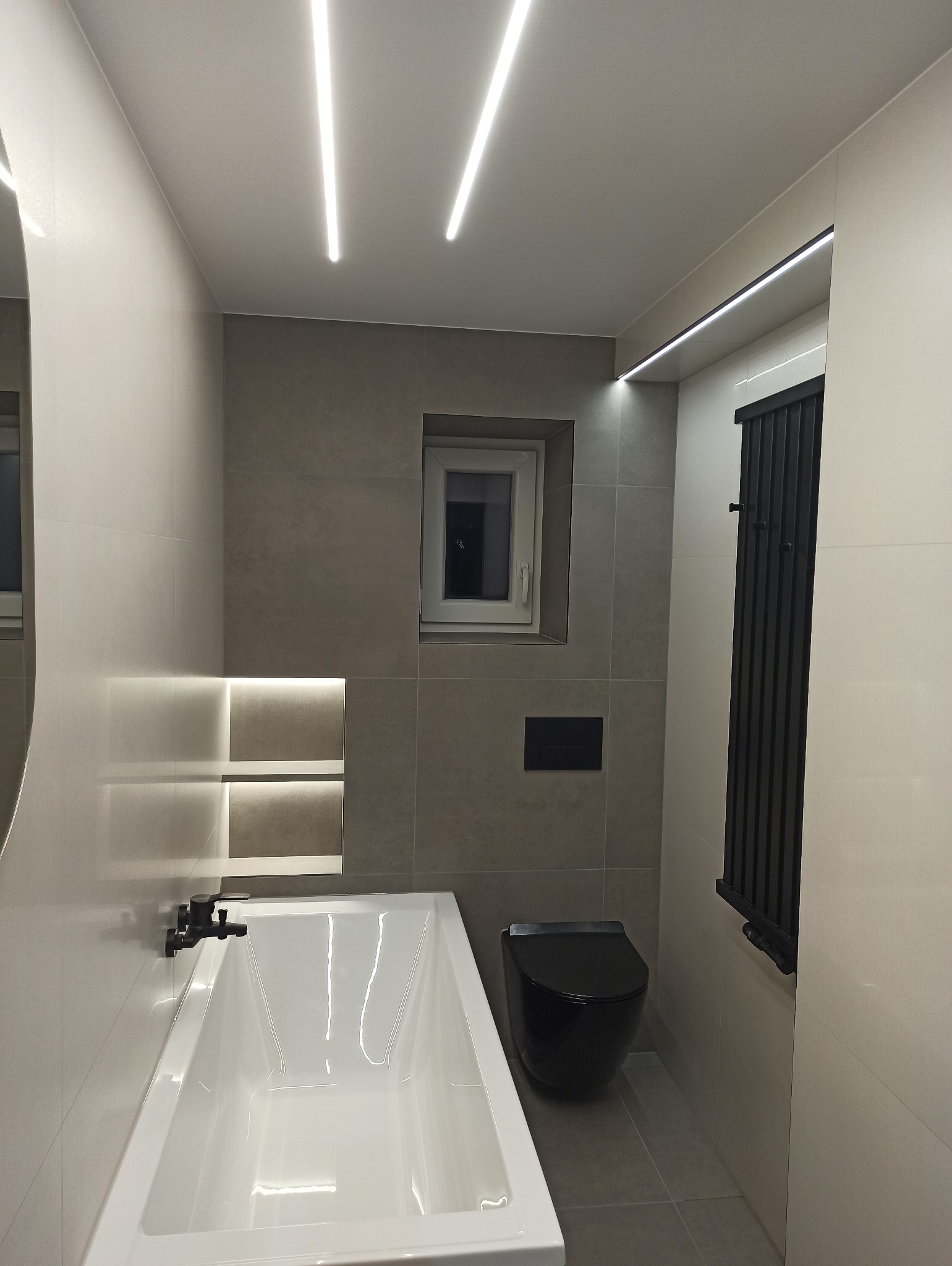 Nowoczesna łazienka z wanną, podwieszaną toaletą w kolorze czarnym i grzejnikiem drabinkowym. Oświetlenie LED w suficie i wnęce nad wanną. Szare płytki.