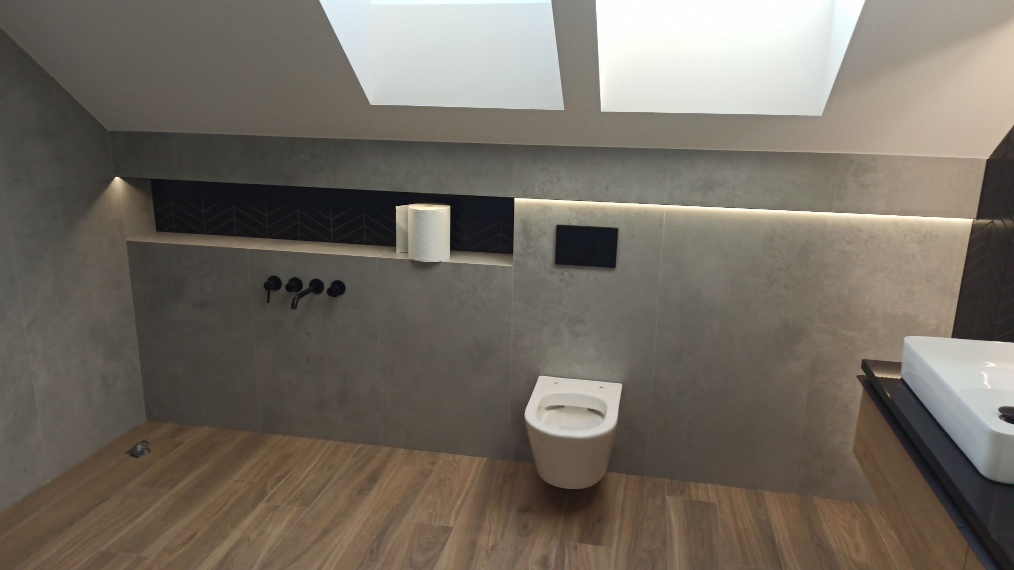 Nowoczesna łazienka na poddaszu z drewnopodobną podłogą, szarą płytką, podświetleniem LED i czarną armaturą. Uwagę zwraca wnęka z dekoracyjną mozaiką.