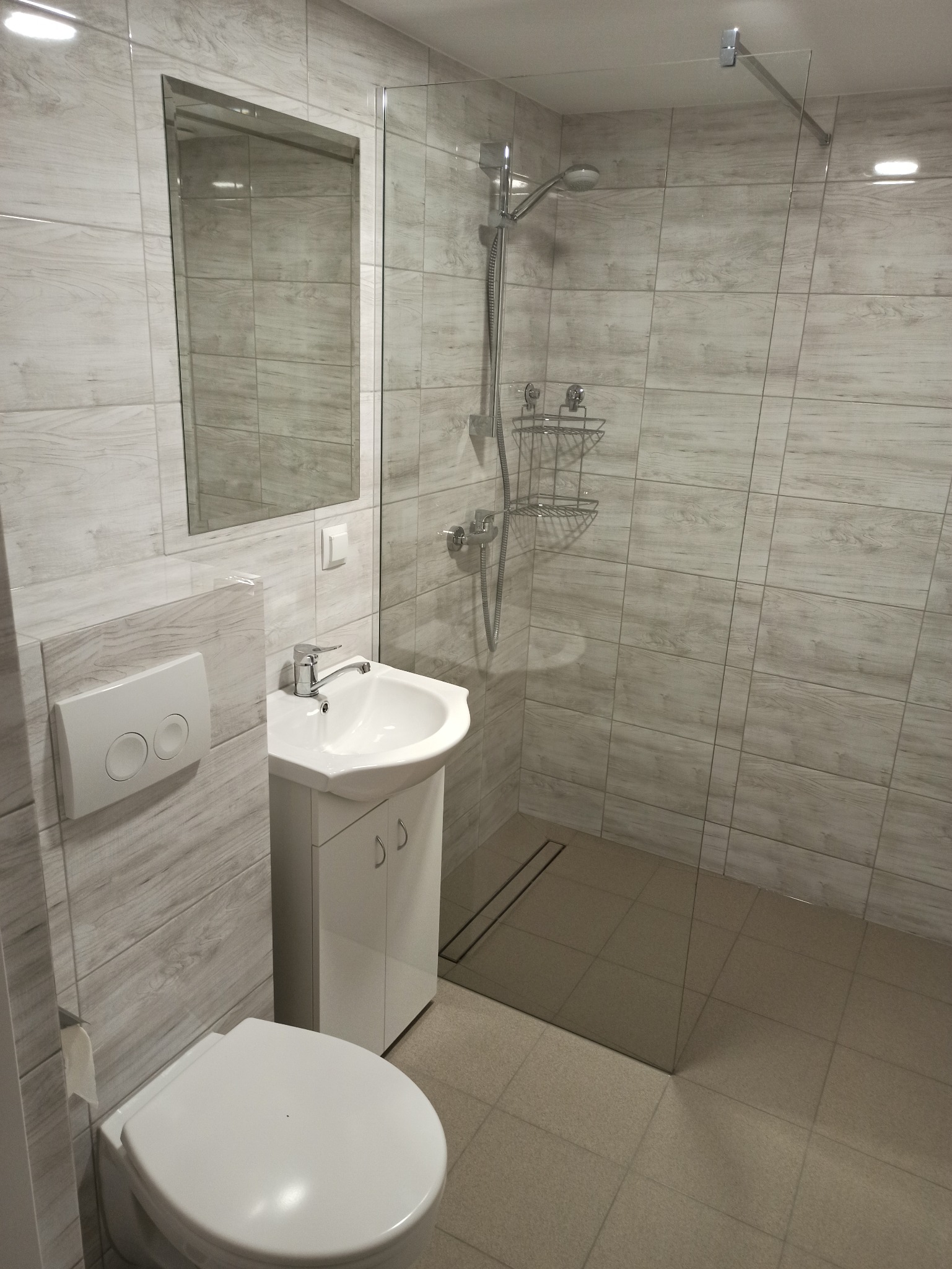 Nowoczesna łazienka z kabiną prysznicową typu walk-in, umywalką z szafką, podwieszaną toaletą i płytkami imitującymi drewno na ścianach.