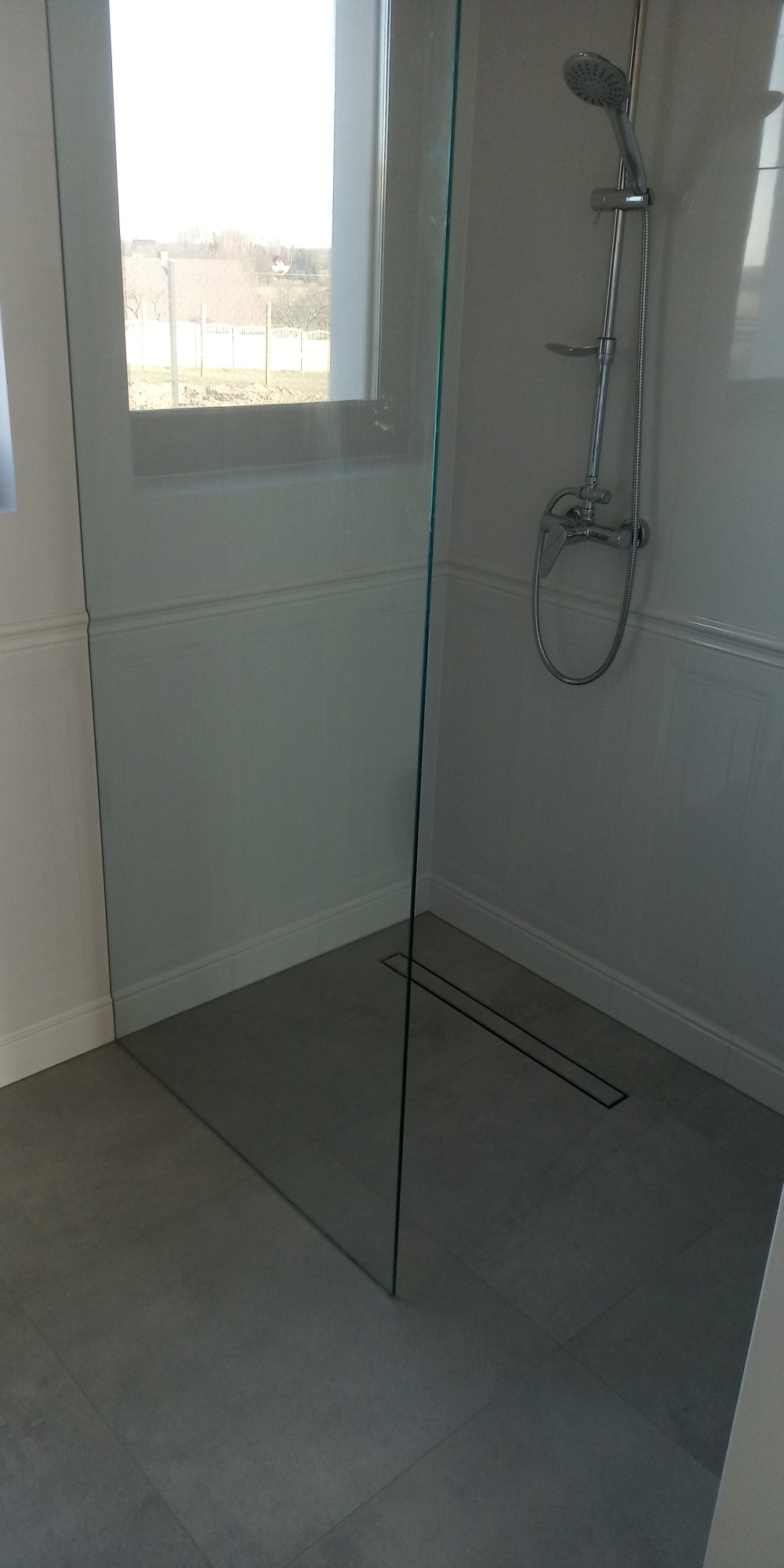 Nowoczesna kabina prysznicowa ze szklaną ścianką, odpływem liniowym w szarej posadzce i widokiem na zewnątrz przez okno.