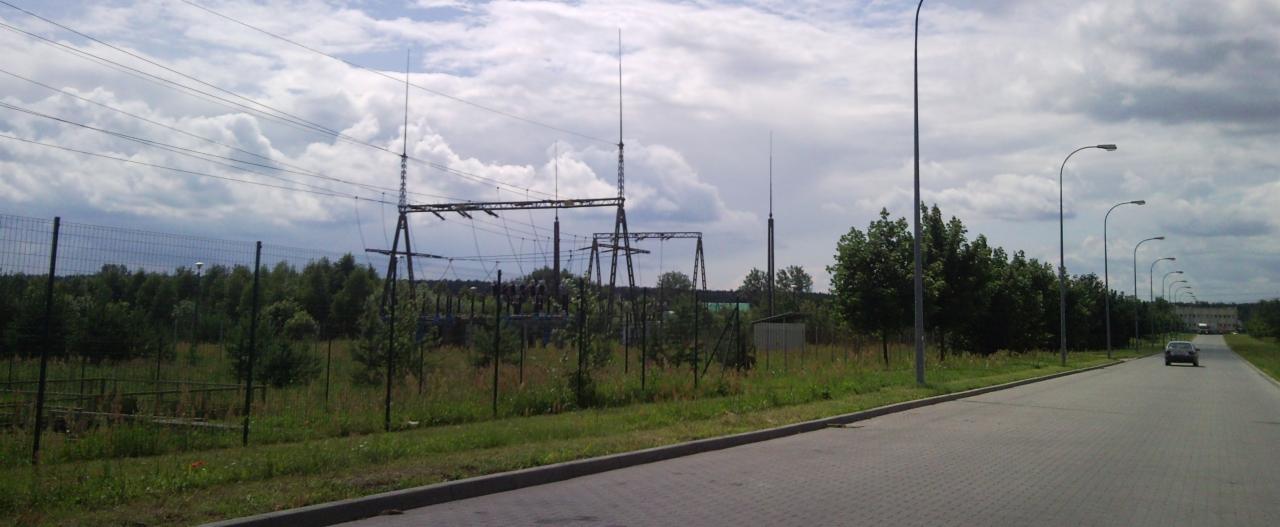 Stacja transformatorowa widziana zza ogrodzenia, z liniami wysokiego napięcia i słupami oświetleniowymi wzdłuż drogi.