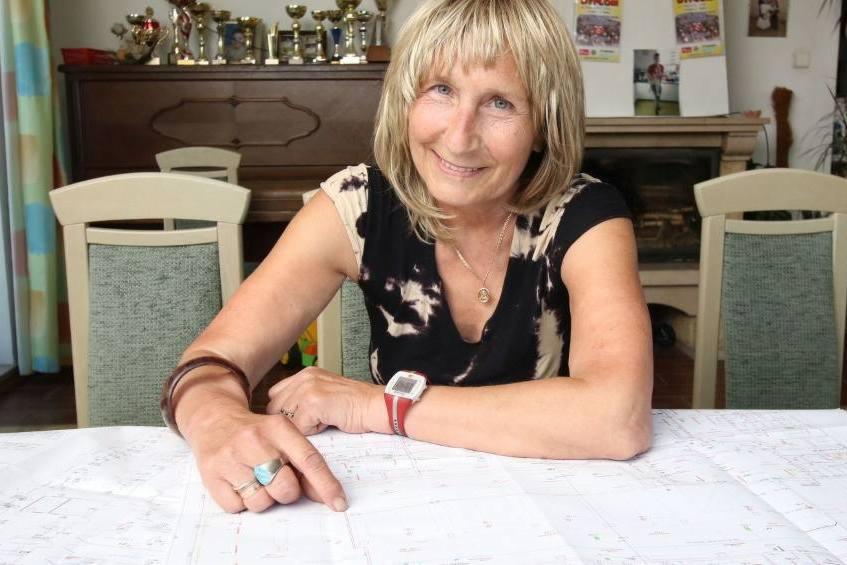 Kobieta z blond włosami wskazuje palcem na plan architektoniczny rozłożony na stole; w tle pianino z trofeami i dyplomami.