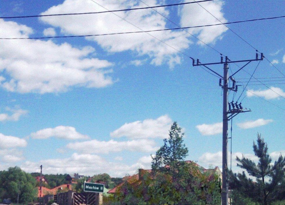 Słup elektryczny z transformatorem na tle błękitnego nieba z obłokami, widoczny znak drogowy z nazwą miejscowości Mastów II.
