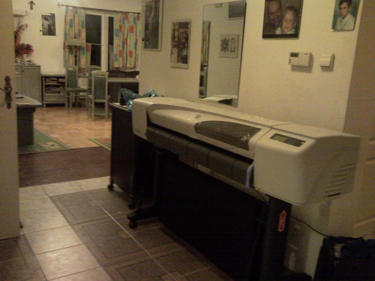 Szerokoformatowa drukarka atramentowa ustawiona w rogu pokoju, widok z boku, na ścianie wiszą obrazy i lustro.