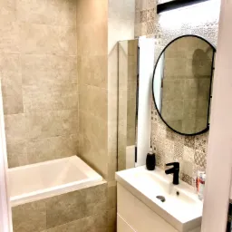 Nowoczesna łazienka z wanną obudowaną szarą płytką, białą szafką z umywalką i czarną armaturą, okrągłym lustrem w czarnej ramie i podłogą imitującą drewno.