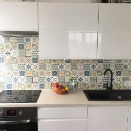 Nowoczesna kuchnia z białymi, błyszczącymi szafkami, mozaikową ścianą nad blatem, czarnym zlewem i płytą indukcyjną, oświetlona naturalnym światłem.