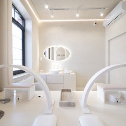 Tofi design - Jasne wnętrze salonu kosmetycznego z dwoma stanowiskami do manicure, oświetlonymi lampami łukowymi z logo 'Afinia', białymi fotelami i frezarkami do paznokci. W tle umywalka z owalnym lustrem...
