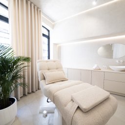 Tofi design - Jasne wnętrze gabinetu kosmetycznego z leżanką z pluszową tapicerką, owalnym lustrem nad umywalką i palmą w białej donicy przy oknie.