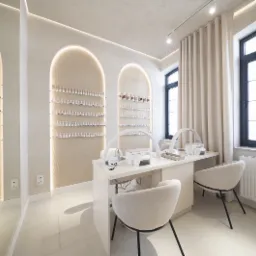 Jasne, minimalistyczne wnętrze salonu kosmetycznego z dwoma stanowiskami do manicure, wyposażonymi w lampy LED i pochłaniacze pyłu, ściany zdobione wnękami z ekspozycją lakierów do paznokci.