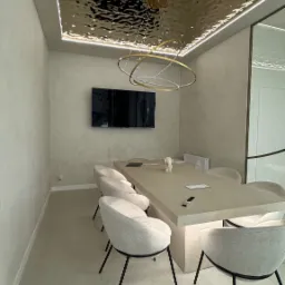 Nowoczesna sala konferencyjna z długim stołem, krzesłami z szarej tkaniny na czarnych nogach, telewizorem na ścianie i dekoracyjnym, lustrzanym sufitem z oświetleniem LED.