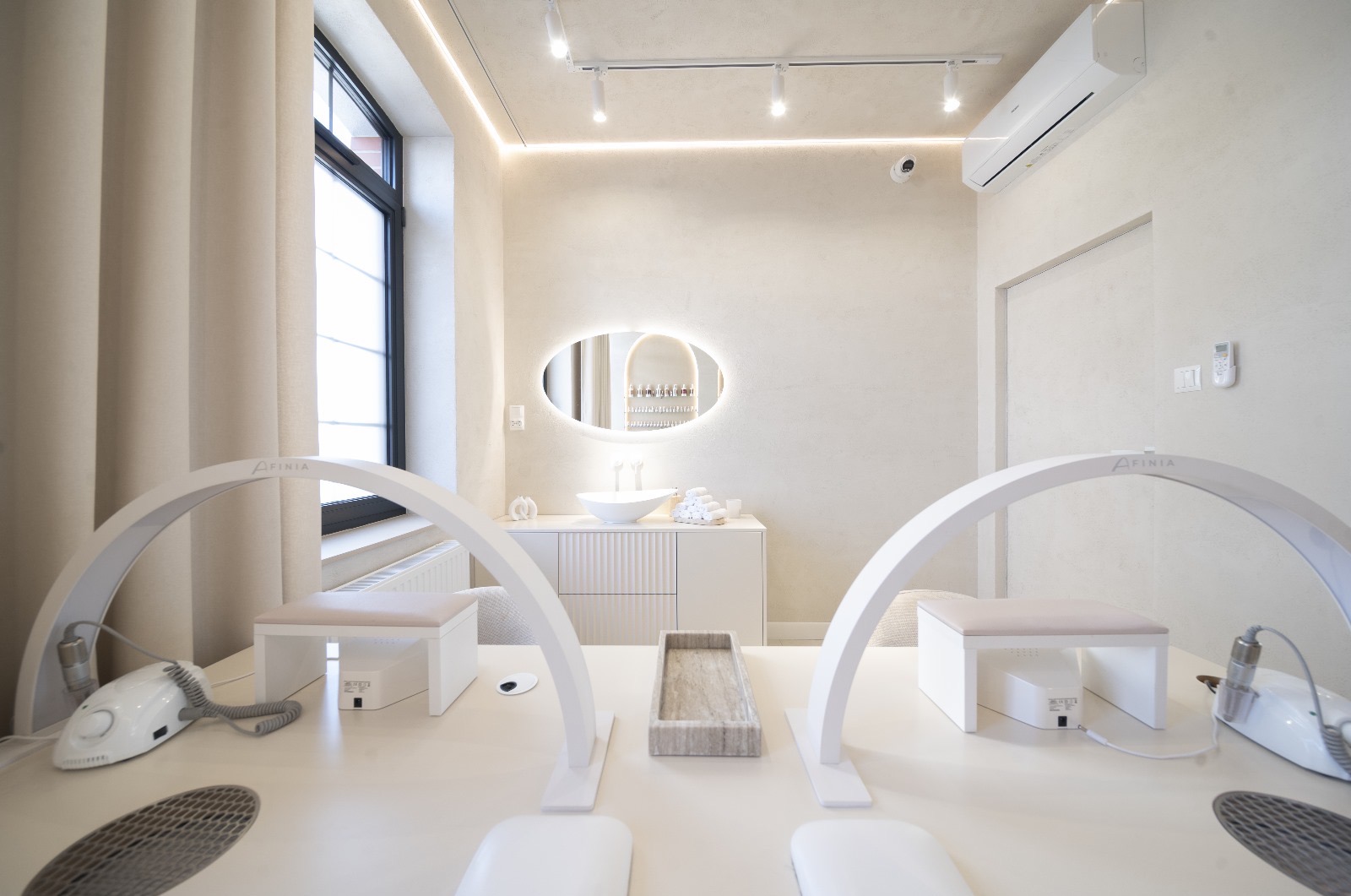 Jasne wnętrze salonu kosmetycznego z dwoma stanowiskami do manicure, oświetlonymi lampami łukowymi z logo 'Afinia', białymi fotelami i frezarkami do paznokci. W tle umywalka z owalnym lustrem...