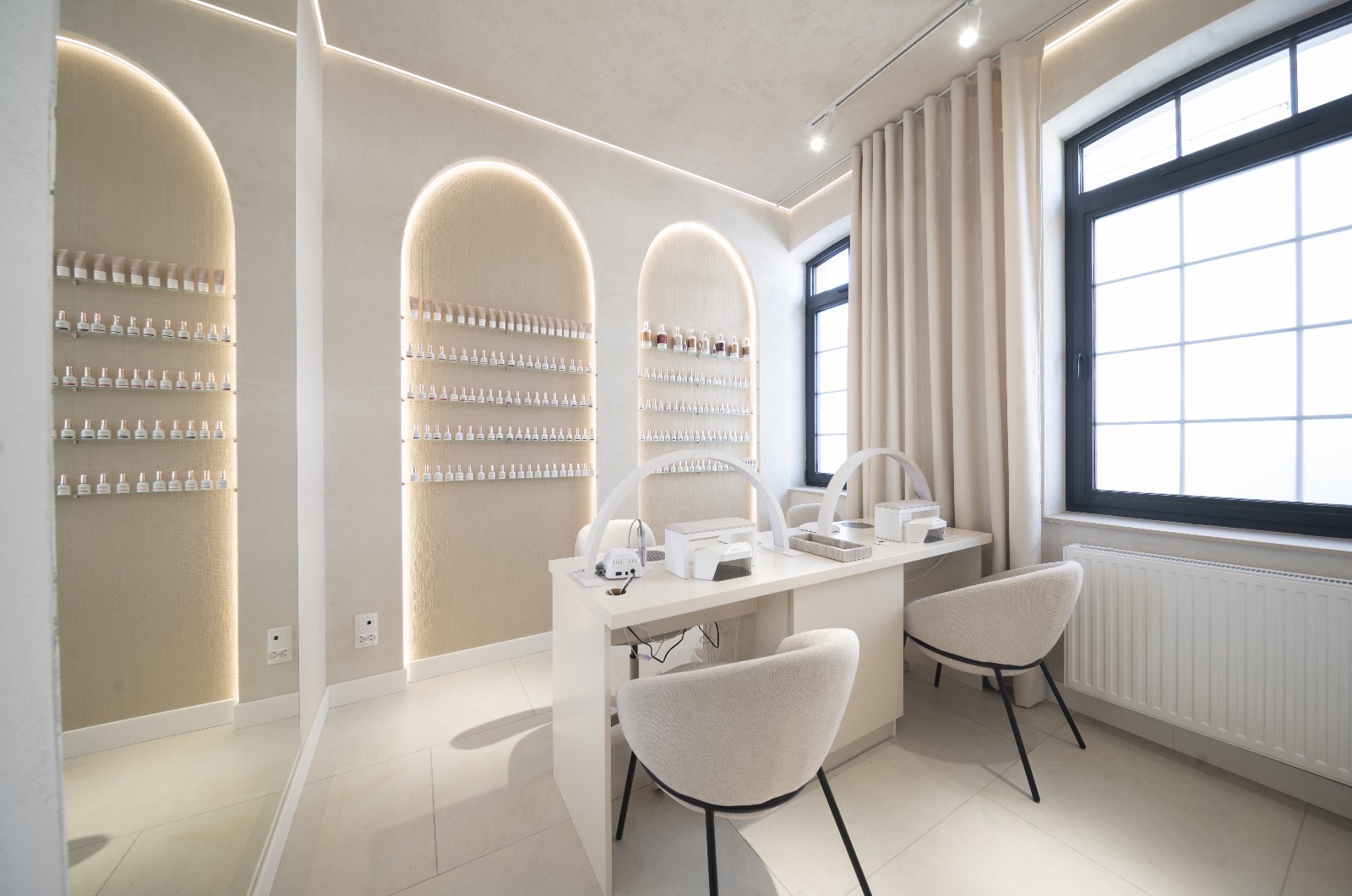 Jasne, minimalistyczne wnętrze salonu kosmetycznego z dwoma stanowiskami do manicure, wyposażonymi w lampy LED i pochłaniacze pyłu, ściany zdobione wnękami z ekspozycją lakierów do paznokci.