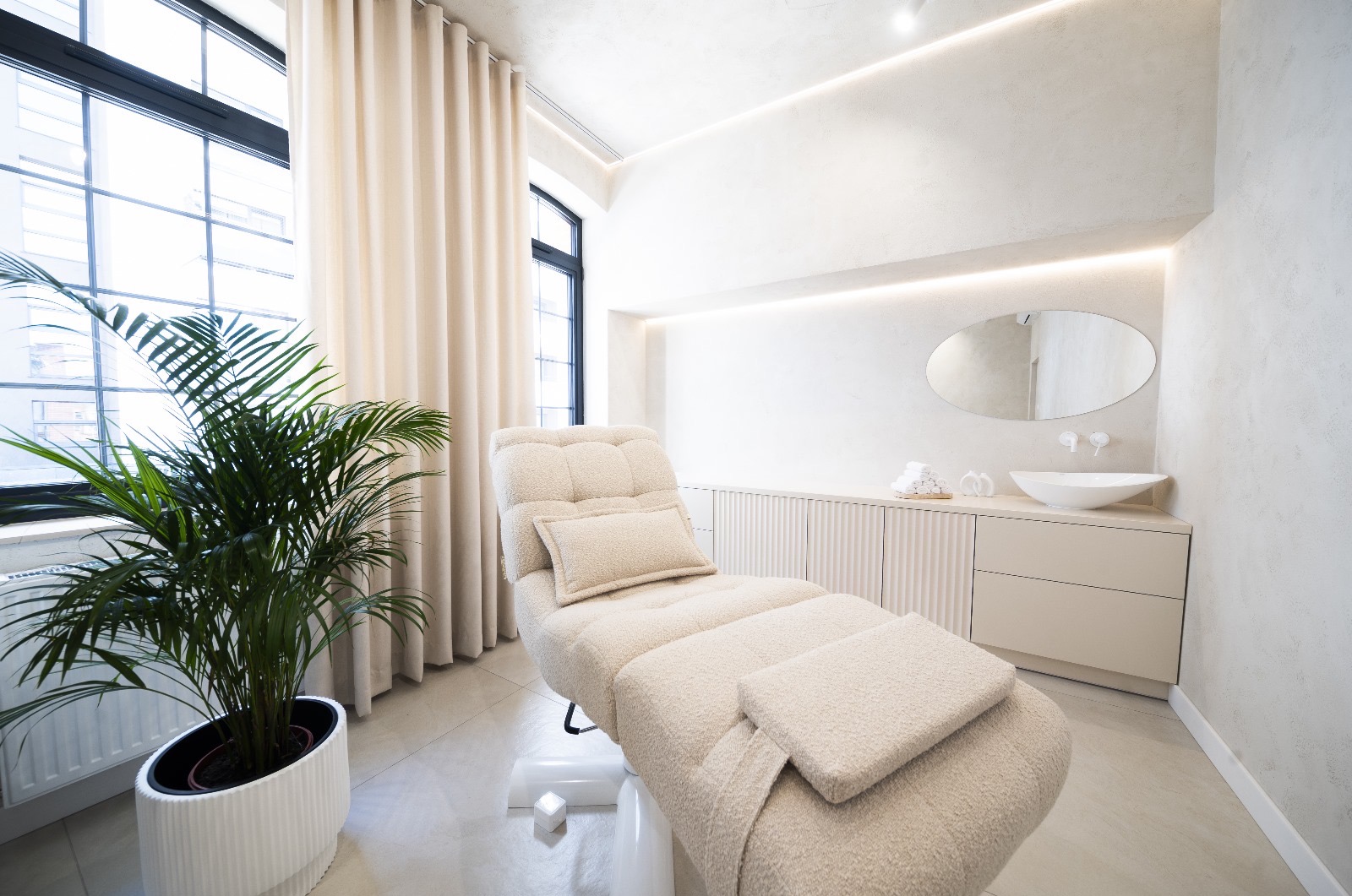 Jasny gabinet kosmetyczny z leżanką obitą tkaniną boucle, owalnym lustrem nad umywalką oraz rośliną w białej donicy przy oknie.