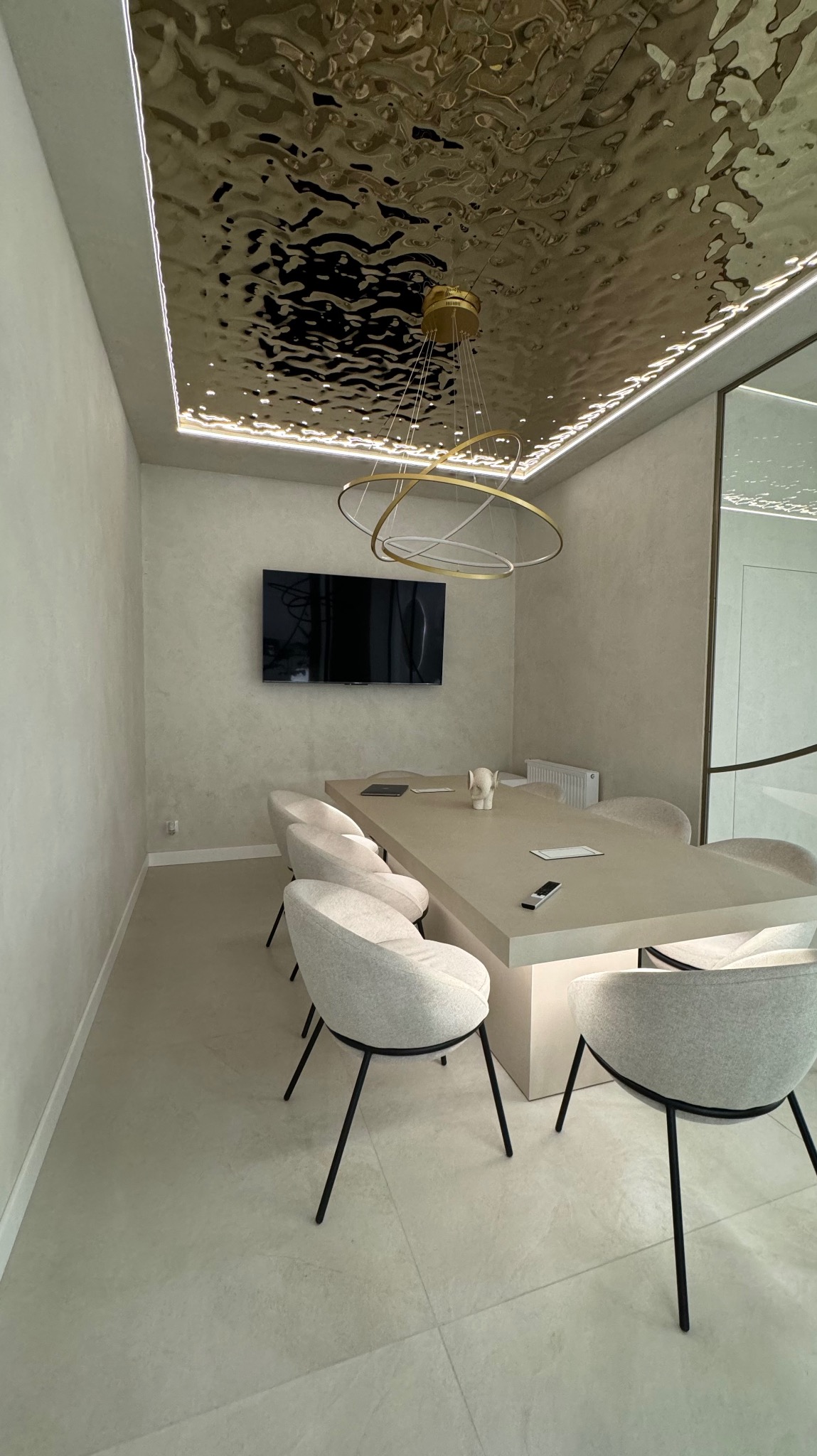 Nowoczesna sala konferencyjna z długim stołem, krzesłami z szarej tkaniny na czarnych nogach, telewizorem na ścianie i dekoracyjnym, lustrzanym sufitem z oświetleniem LED.