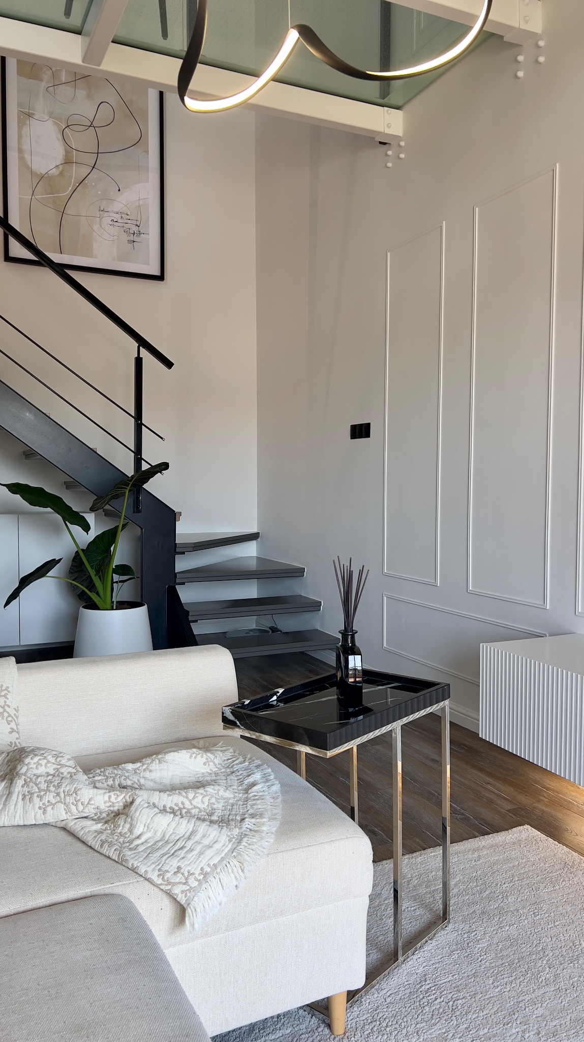 Jasne wnętrze salonu z minimalistycznymi schodami, rośliną w białej donicy, beżową sofą z dekoracyjnym pledem, czarnym stolikiem pomocniczym z marmurowym blatem i designerską lampą sufitową.