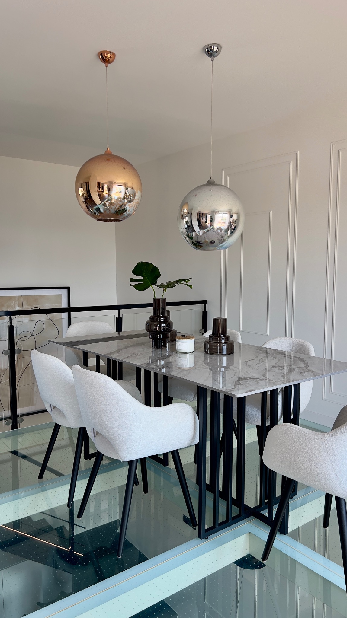 Jadalnia z marmurowym stołem, białymi krzesłami na czarnych nogach i szklaną podłogą, oświetlona dwoma kulistymi lampami: miedzianą i srebrną.