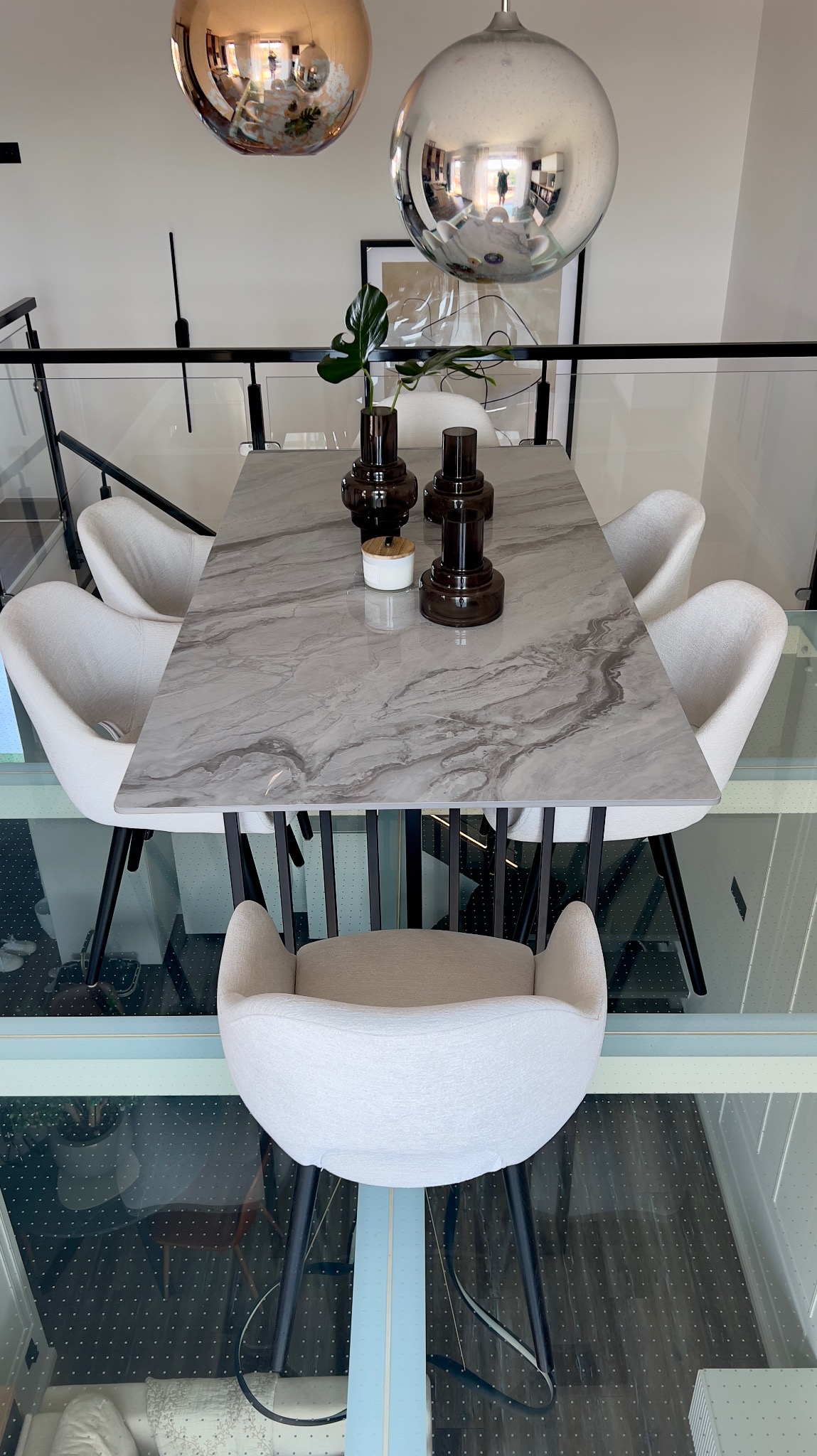 Marmurowy stół jadalny z białymi krzesłami na tle szklanej podłogi, dekorowany wazonami i odbijającymi światło kulistymi lampami wiszącymi.