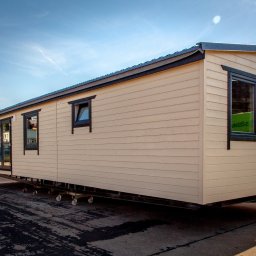 Przedsiębiorstwo Partner Sp. z o.o. - Gotowy dom modułowy z jasną elewacją i ciemnymi ramami okiennymi, ustawiony na platformie transportowej. Widoczne ślady opon na placu.