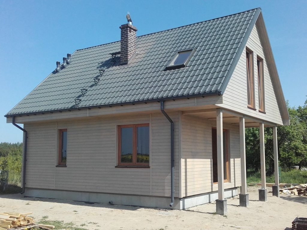 Gotowy dom szkieletowy z szarą dachówką i drewnianą elewacją, z widocznym kominem z cegły i elementami wykończeniowymi. Jasne, słoneczne ujęcie.