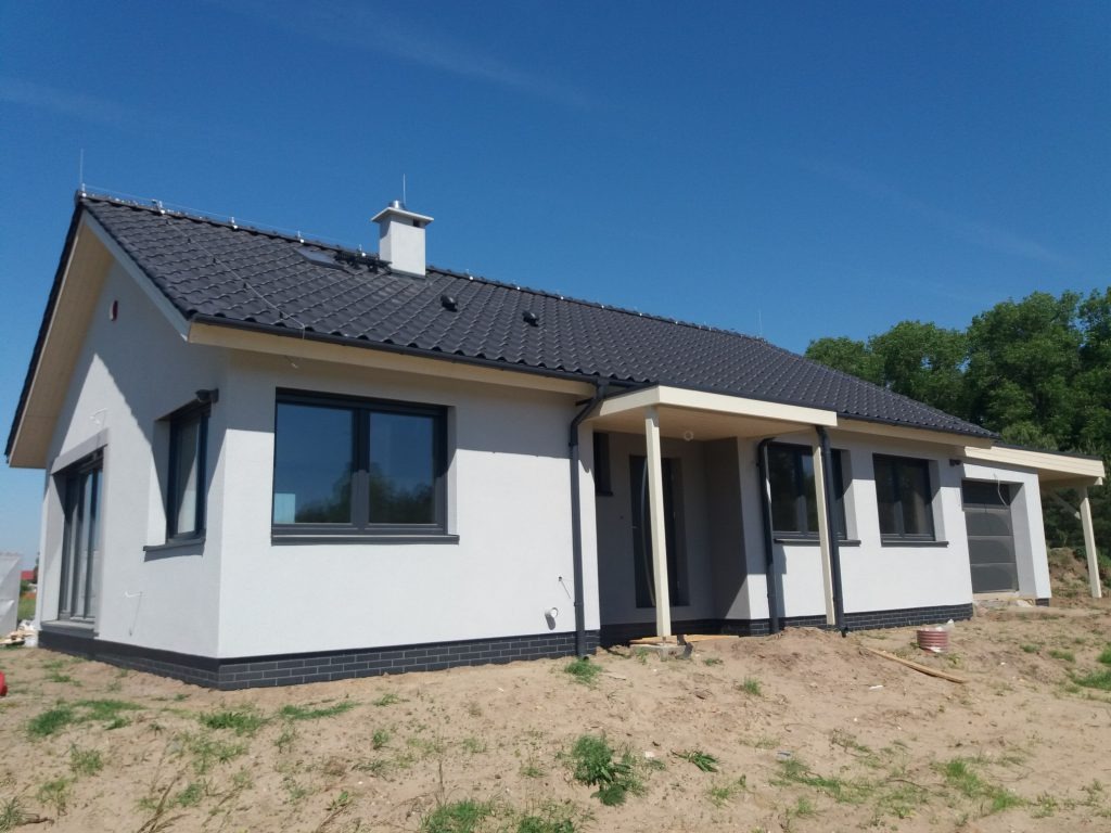 Parterowy dom z ciemną dachówką i jasną elewacją, zadaszone wejście i garaż, widok z zewnątrz w słoneczny dzień. Detal: czarny pas cegieł przy fundamencie.