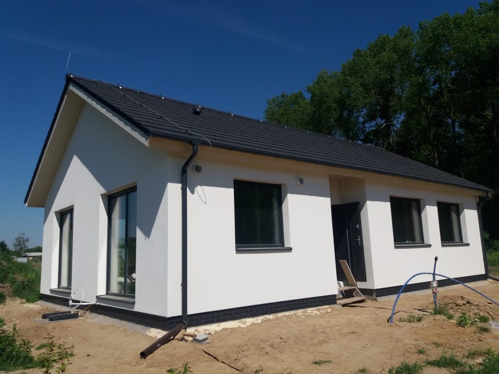 Biały, parterowy dom z ciemną dachówką i czarnymi oknami, widziany z zewnątrz w słoneczny dzień. Widoczne elementy wykończeniowe i fragment terenu.