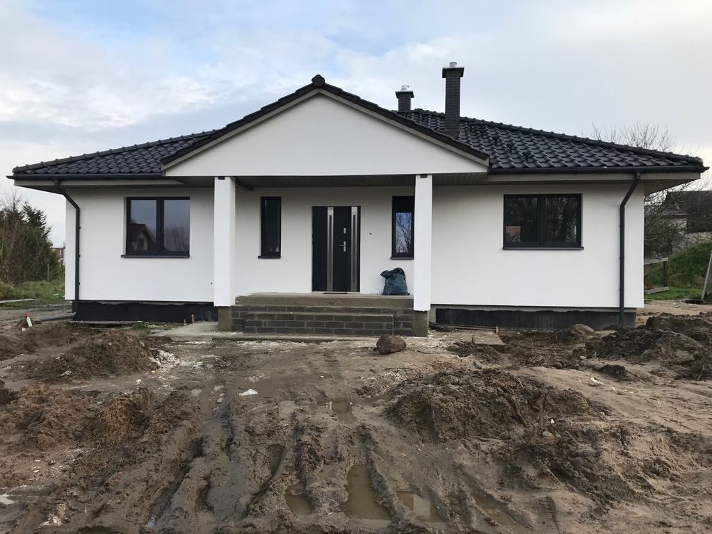 Biały dom z czarnym dachem i oknami, widok z przodu na błotnistej działce. Widoczne schody wejściowe i elementy wykończeniowe.