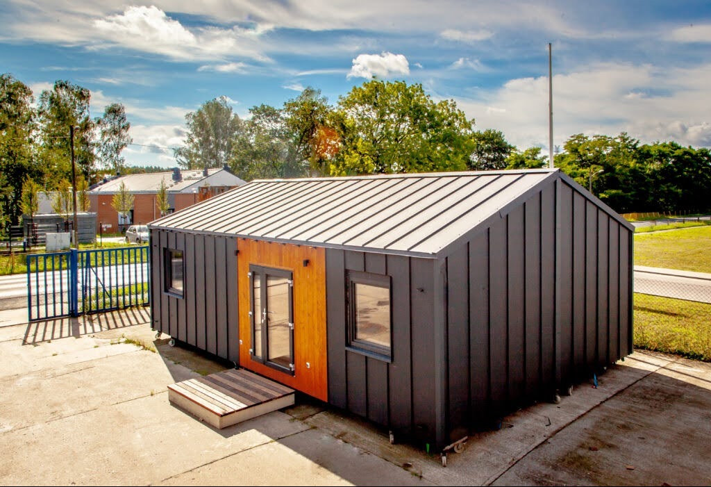 Gotowy, czarny dom modułowy z drewnianymi akcentami, widoczny z zewnątrz na betonowej powierzchni, z zielonym tłem drzew i błękitnym niebem.