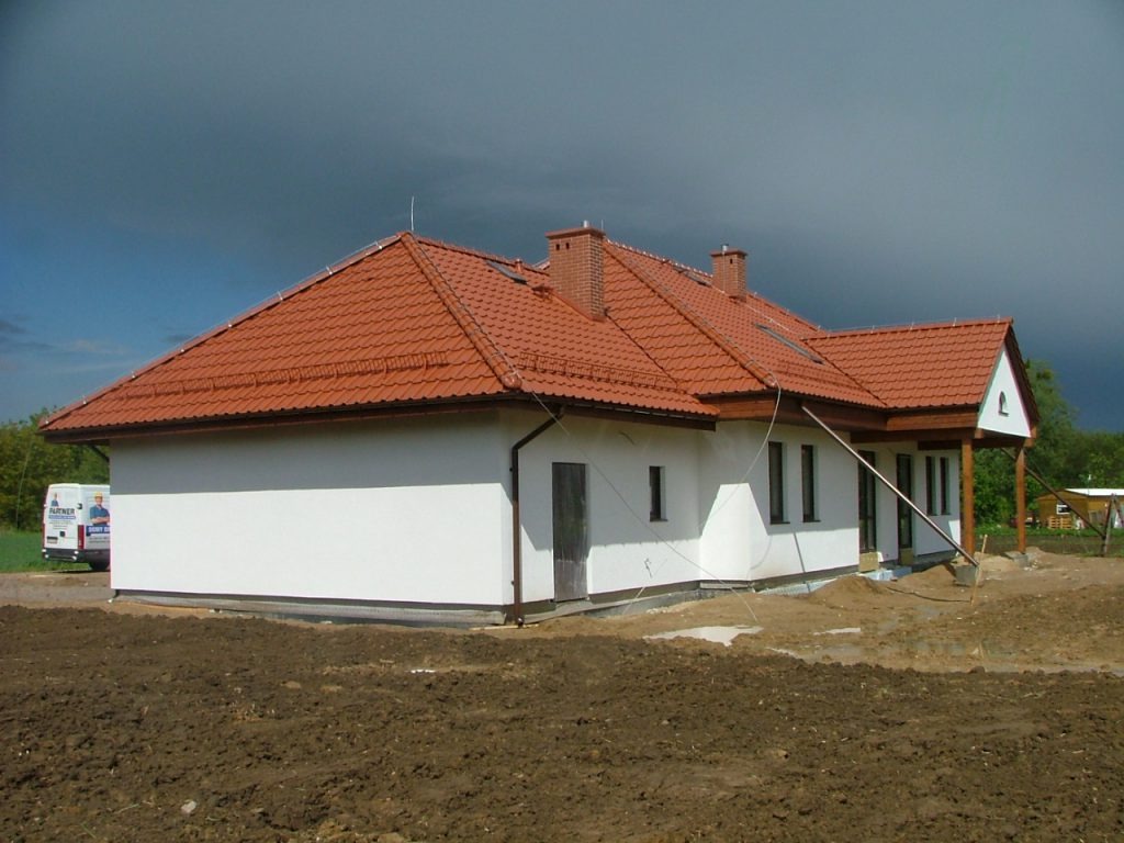 Biały dom z czerwoną dachówką w trakcie budowy, widoczny fragment ogrodu i pochmurne niebo. Na posesji stoi biały bus.