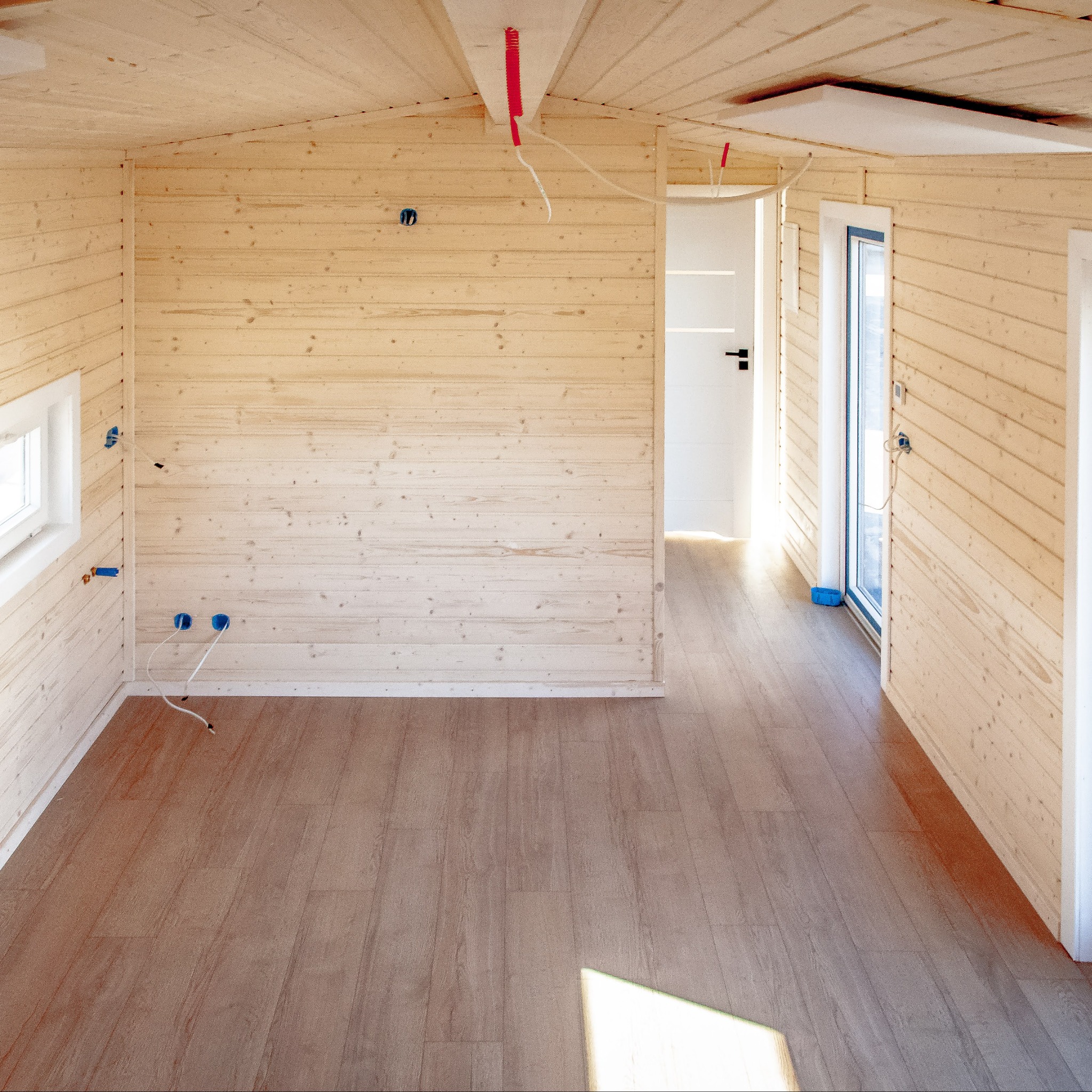 Wnętrze domu szkieletowego z drewnianą boazerią i panelami podłogowymi. Widoczne instalacje elektryczne i okno. Jasne, naturalne światło.