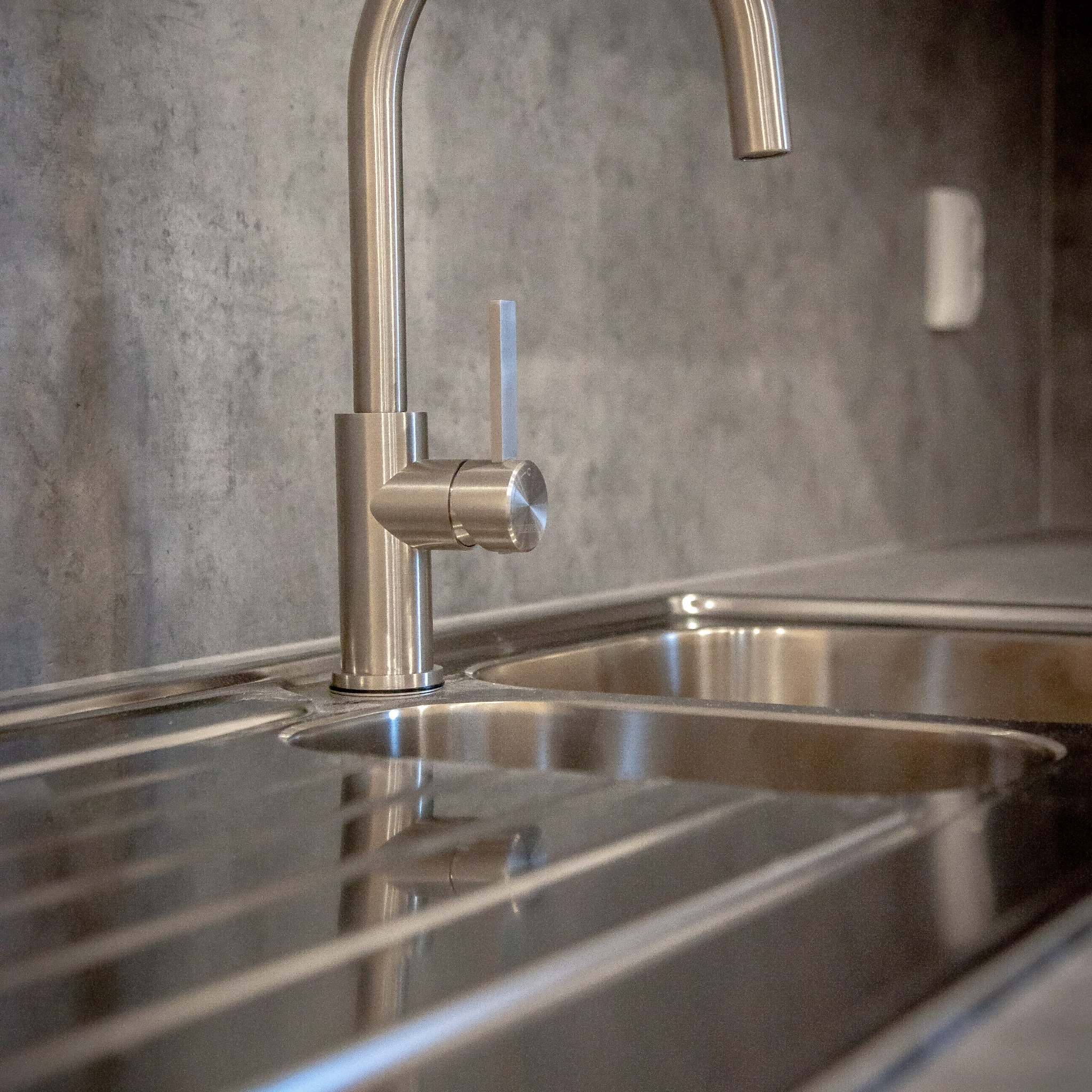 Błyszcząca, metalowa bateria kuchenna z pojedynczym uchwytem, zamontowana na dwukomorowym zlewozmywaku ze stali nierdzewnej na tle szarej ściany.