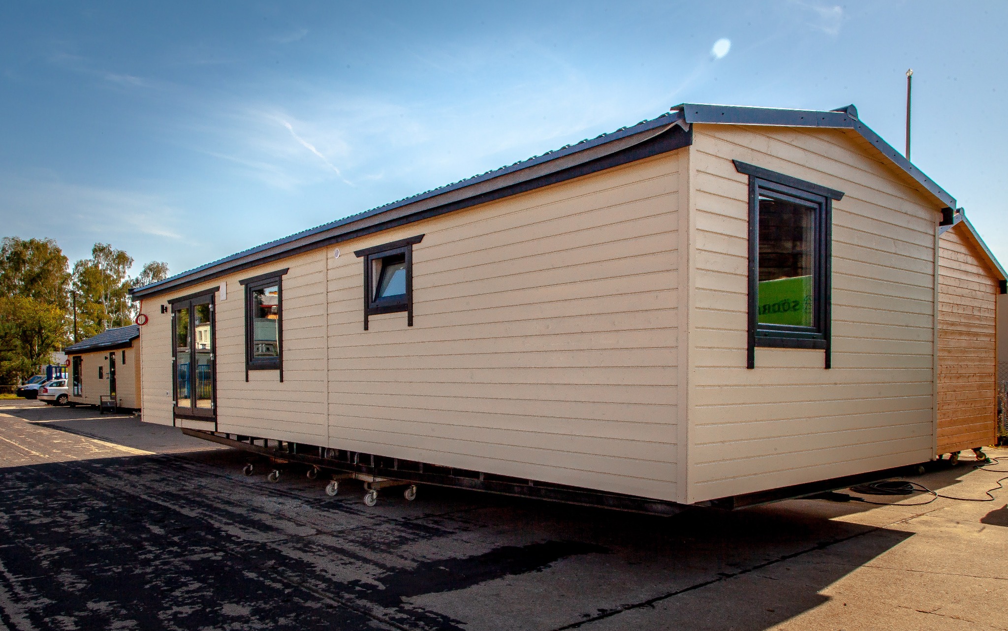 Gotowy dom modułowy z jasną elewacją i ciemnymi ramami okiennymi, ustawiony na platformie transportowej. Widoczne ślady opon na placu.