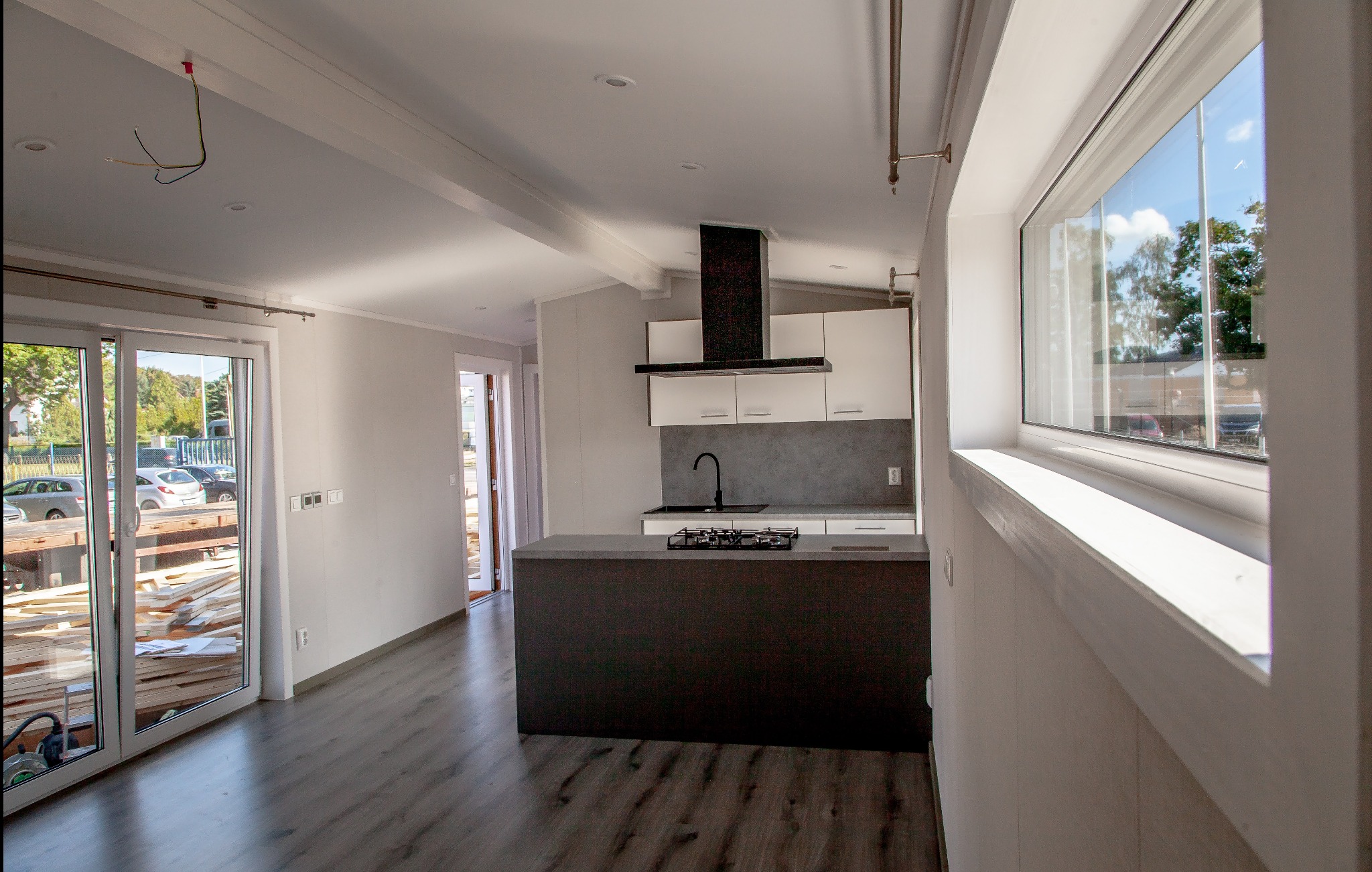 Jasne wnętrze domu modułowego z widoczną kuchnią i stertą drewna przy przeszklonych drzwiach tarasowych. Minimalistyczny design, surowe wykończenie podłogi.