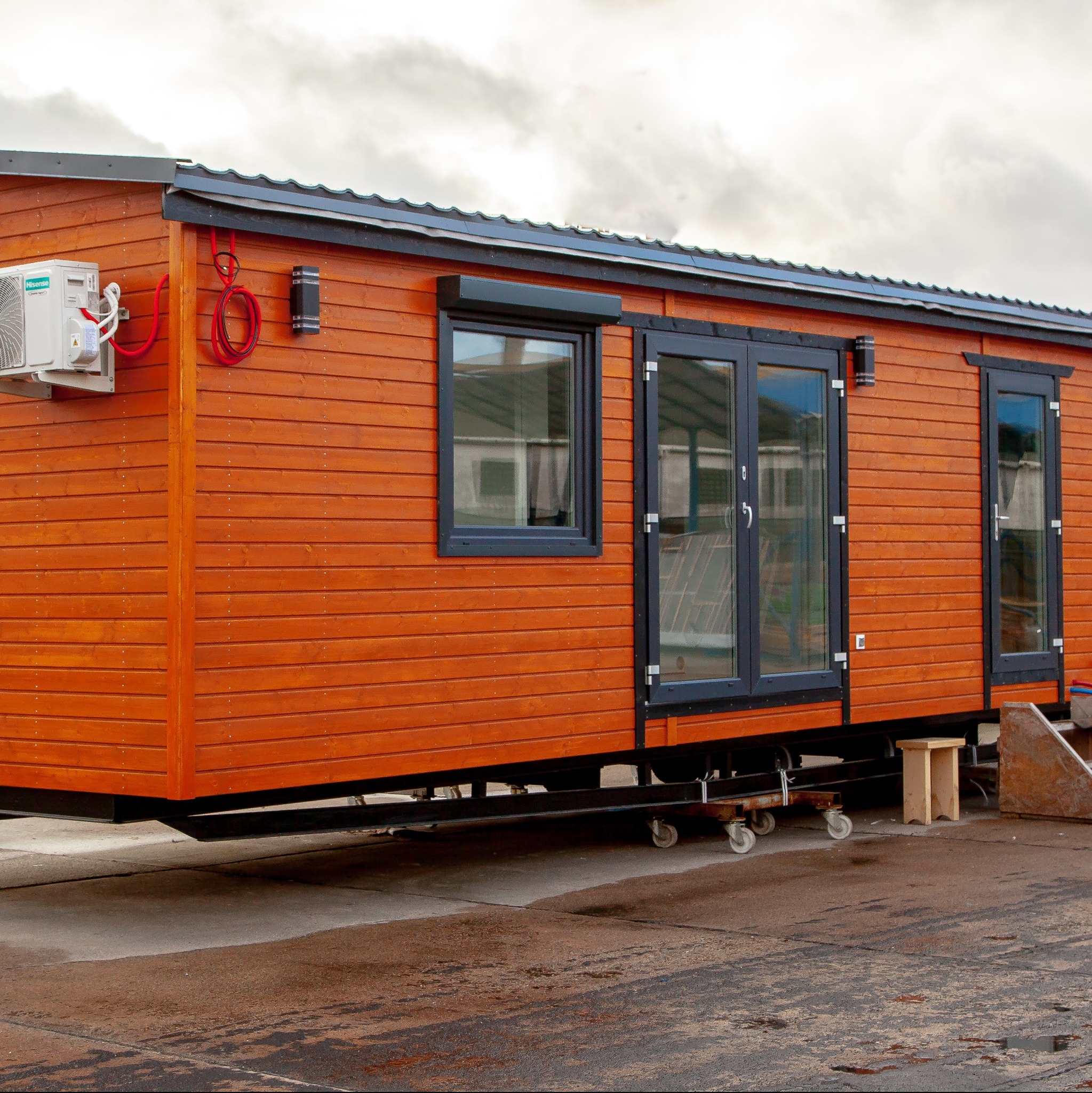 Nowoczesny dom modułowy z drewnianą elewacją, czarnymi ramami okien i klimatyzacją na zewnątrz, osadzony na mobilnej platformie z kołami.