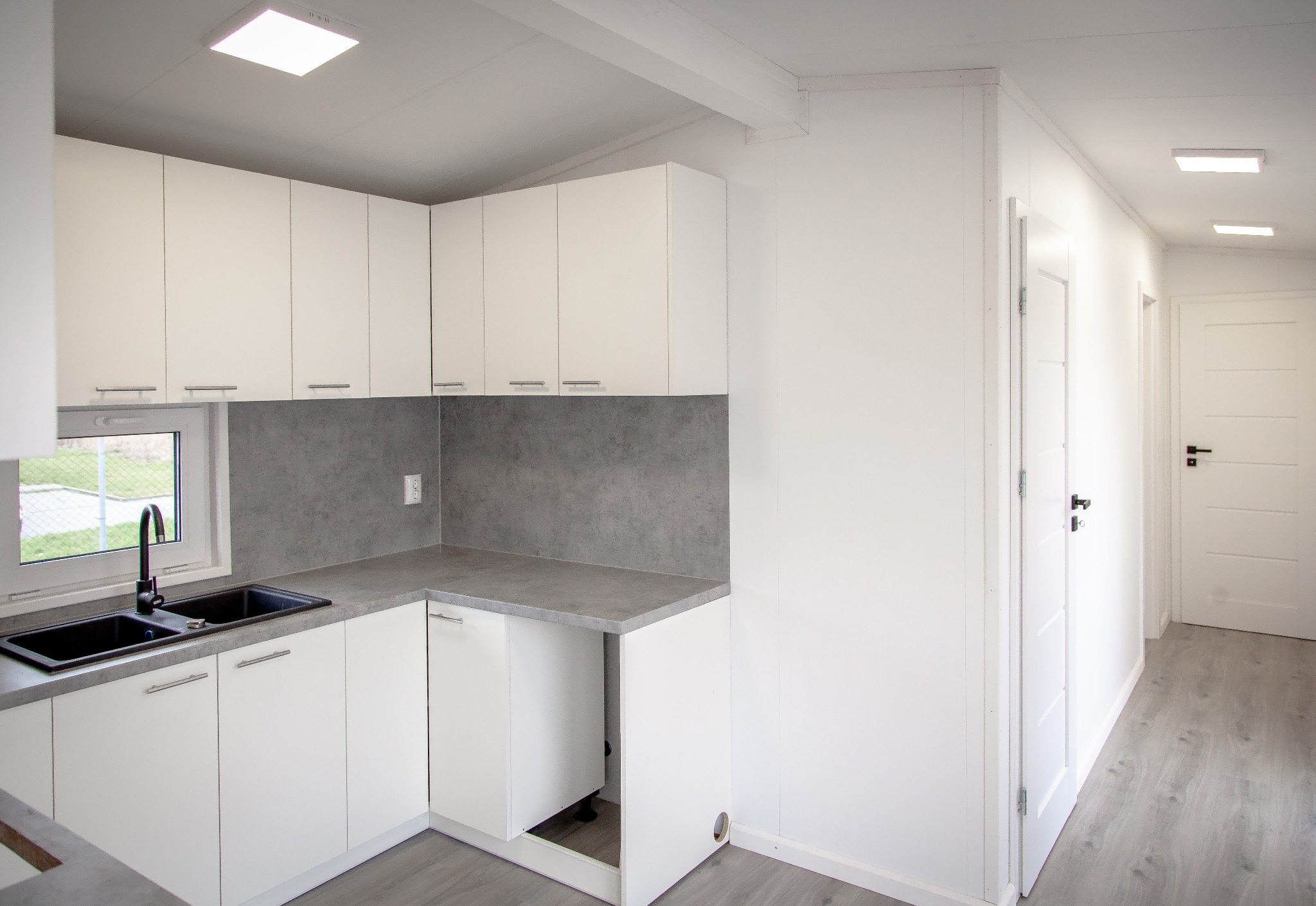 Wnętrze modułowego domu: minimalistyczna kuchnia z białymi szafkami i szarym blatem, widok na korytarz z białymi drzwiami i szarą podłogą.