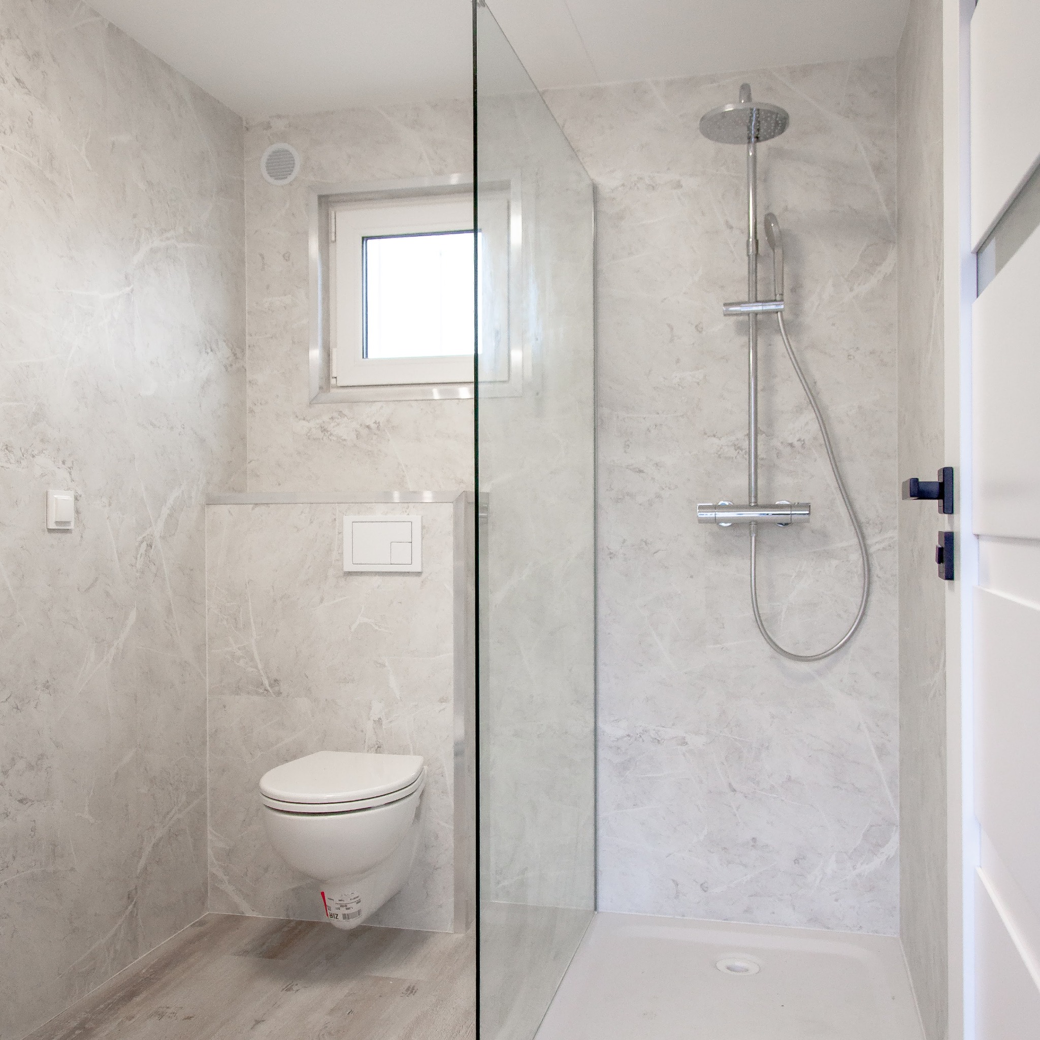 Nowoczesna łazienka z kabiną prysznicową typu walk-in, toaletą podwieszaną i szarą marmurową okładziną ścian. Widoczne białe drzwi i okno.