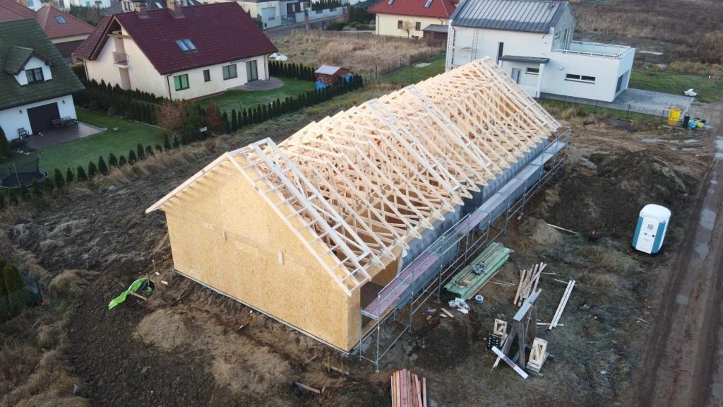 Wznoszenie konstrukcji domu szkieletowego z drewnianym szkieletem dachu, widok z góry na plac budowy z rusztowaniami i przenośną toaletą.