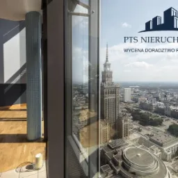 Wnętrze apartamentu z widokiem na Pałac Kultury i Nauki w Warszawie, kolumna wykończona mozaiką, drewniana podłoga, logo PTS Nieruchomości widoczne w odbiciu okna.