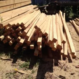 Stos świeżo przetartego drewna o przekroju kwadratowym, ułożony na ziemi w pobliżu ściany budynku.
