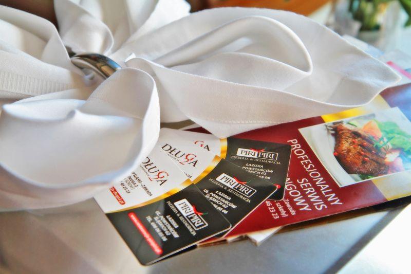 Elegancko złożona biała serwetka z pierścieniem na wizytówkach i ulotkach restauracji Piri Piri i Długa, z widocznym zdjęciem dania mięsnego.