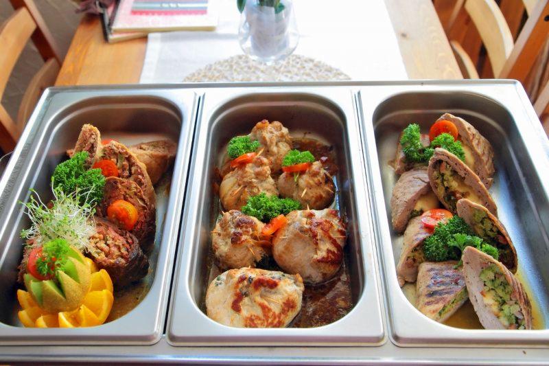 Trzy metalowe pojemniki GN z różnymi rodzajami mięsnych roladek, ozdobione warzywami i pomidorkami koktajlowymi, ustawione na drewnianym stole.