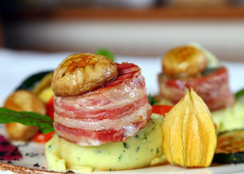 Dwa medaliony z polędwicy owinięte boczkiem, zwieńczone pieczarkami i podane na puree ziemniaczanym z dodatkiem warzyw i miechunki peruwiańskiej.