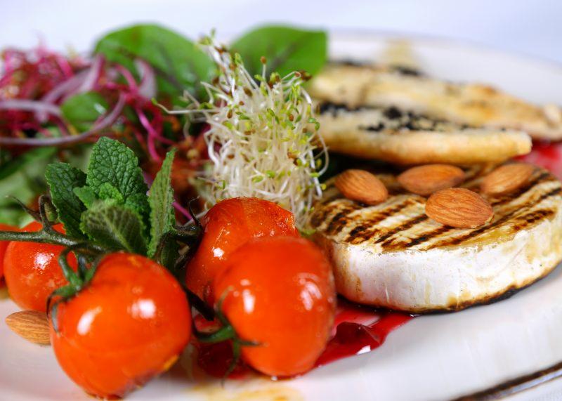 Grillowany ser camembert z migdałami, pomidorkami cherry, kiełkami i mieszanką sałat na białym talerzu, polany sosem.