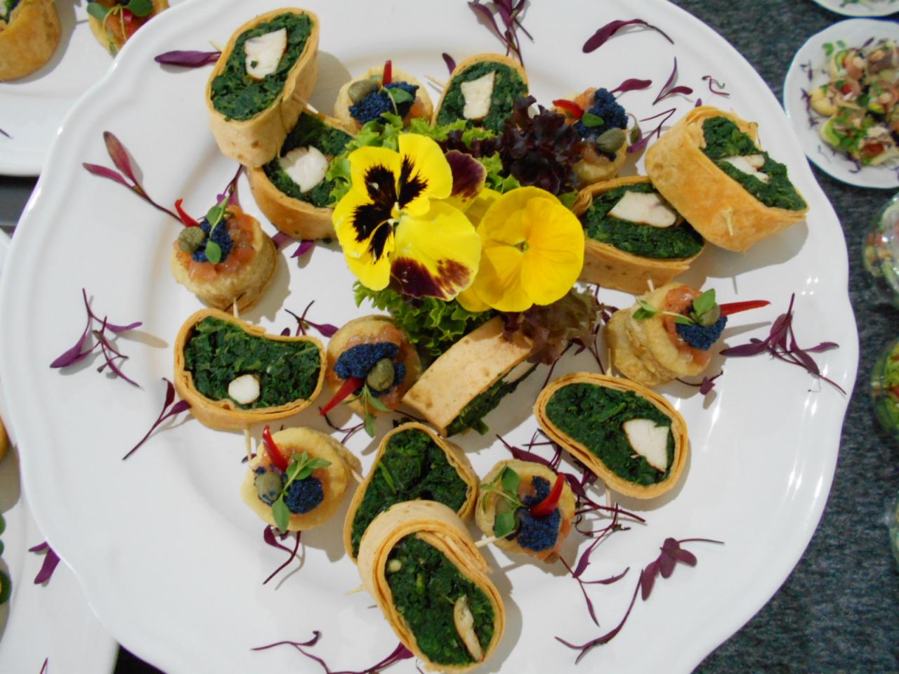 Elegancko ułożone na białym talerzu mini przekąski: roladki z ciasta z nadzieniem szpinakowym i kawałkiem sera, oraz tartaletki z łososiem i kawiorem, udekorowane jadalnymi kwiatami i purpurowymi...