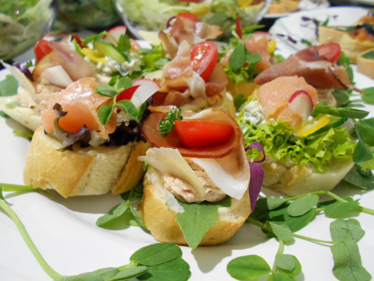 Dekoracyjne kanapeczki na białym talerzu, z łososiem, szynką parmeńską, pomidorkami cherry, rzodkiewką i świeżymi ziołami, podane na jasnym pieczywie.
