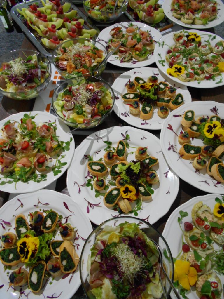 Stół cateringowy z różnorodnymi przekąskami: sałatki w szklanych misach, kanapeczki z pastą szpinakową i jadalnymi kwiatami na białych talerzach z motywem kwiatowym, kompozycje z melona, truskawek...