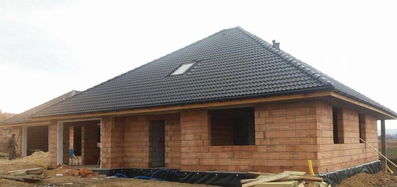 Dom w trakcie budowy z ciemną dachówką i oknem dachowym, widoczne bloczki z czerwonej cegły, czarna folia fundamentowa i składowane drewno