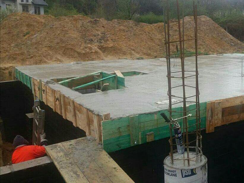 Wylewka fundamentów z szalunkiem i zbrojeniami pionowymi, widoczny robotnik w wykopie.