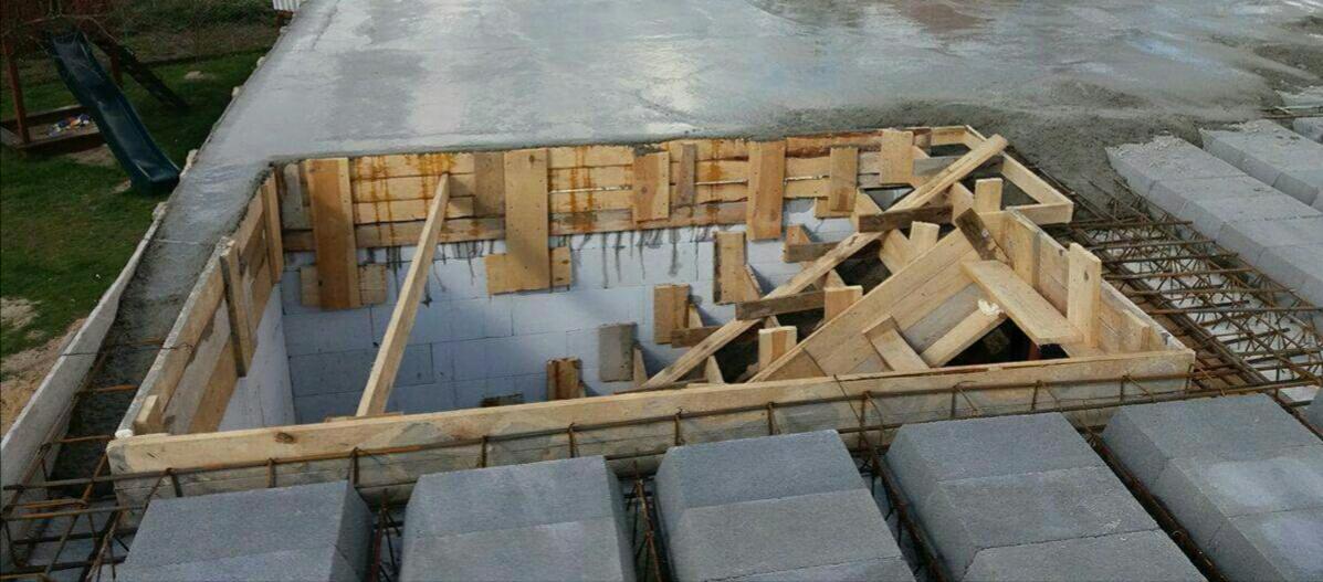 Wykop pod fundamenty z drewnianym szalunkiem, zbrojony stalową siatką i częściowo zalany betonem, widoczne bloczki betonowe i fragment placu zabaw w tle.